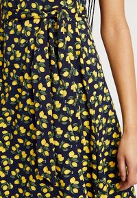 Marinblå klänning med ett mönster av citroner och blad, med knyt i midjan, veckade detaljer och en mjuk tygstruktur.