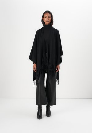 Svart poncho i mjukt tyg, med lös passform, spetsig nederkant och fransade kanter. Matchad med vida svarta byxor och klackboots.