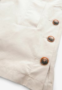 Tissu beige avec trois boutons ronds marron et gris alignés verticalement près d'une poche et des coutures cousues.
