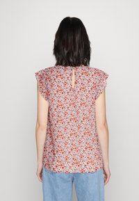 Frau mit schulterlangem dunklem Haar, die ein ärmelloses Blumenoberteil mit roten und lila Blumen sowie eine hellblaue Jeans trägt, von hinten gesehen.