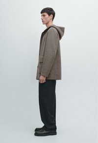 Cappotto marrone con cappuccio in una miscela di lana strutturata, con tasche laterali e chiusura a bottoni. Abbinato a pantaloni neri a gamba larga e scarpe.