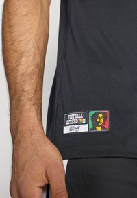 Μαύρο αθλητικό πουκάμισο με απαλό ύφασμα, με ένα σχέδιο που γράφει «TO FOOTBALL ΕΙΝΑΙ ΕΛΕΥΘΕΡΙΑ» δίπλα σε πολύχρωμο πορτρέτο του Μπομπ Μάρλεϊ.