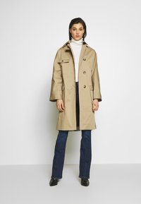 Beige trenchcoat met een knoopsluiting aan de voorkant, grote zakken en een kraag, gecombineerd met blauwe jeans en zwarte laarzen. Soepele textuur, klassiek ontwerp.