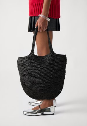Sac fourre-tout noir tissé de forme ronde, en matériau texturé, avec des poignées doubles. Sans embellissements, présenté tenu par une personne.