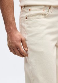 Ανδρικό χέρι που ακουμπάει δίπλα σε ανοιχτό μπεζ τζιν από ντένιμ με ορατή ραφή και χαλκομανίες σε απλό φόντο.