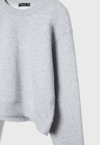 Sweatshirt à col rond à manches longues gris clair, avec des poignets et un ourlet côtelés, avec une étiquette de marque visible à l'intérieur du col.