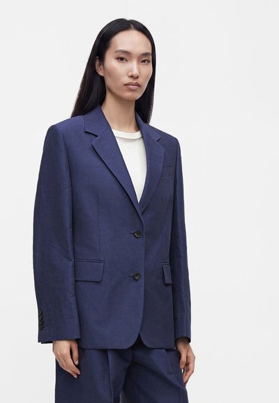 Femme aux longs cheveux foncés portant un blazer bleu marine et un pantalon assorti sur un haut blanc, sur un fond clair uni.