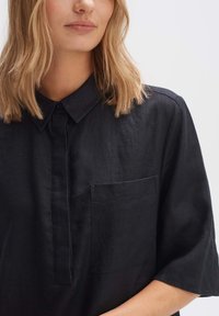 Camicia nera in lino con colletto button-down, maniche corte e tasca sul petto. Il tessuto ha una finitura texturizzata.
