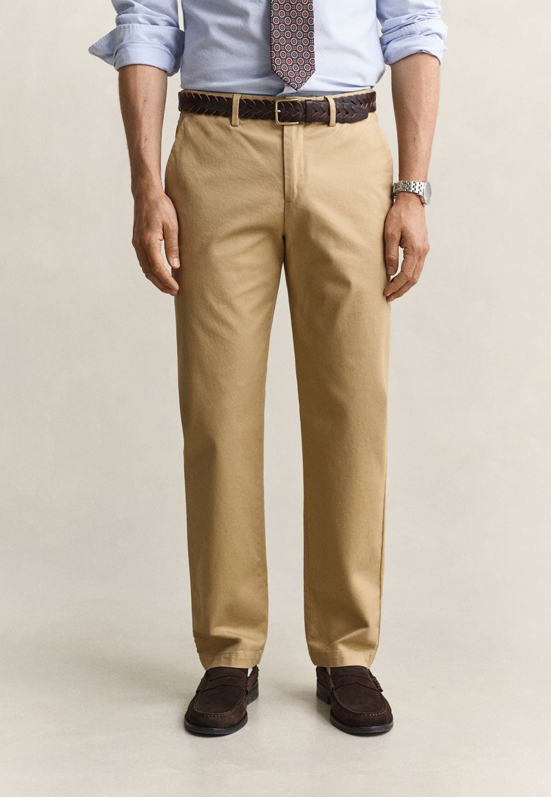 Homme portant un pantalon beige, une ceinture tressée marron, une chemise bleu clair retroussée, une cravate à motifs, une montre argentée et des mocassins marron foncé.