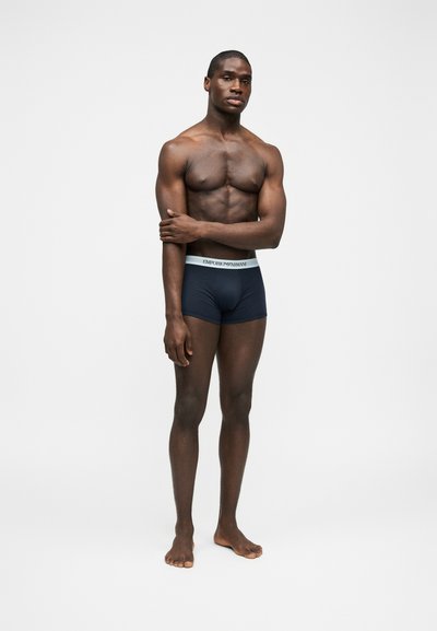 Mężczyzna o ciemnej karnacji i umięśnionej sylwetce, stojący boso w granatowych bokserkach Emporio Armani, trzymający jedną rękę na klatce piersiowej na jednolitym tle.