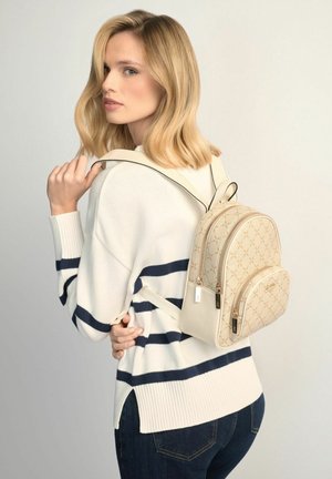 Blondynka w białym swetrze z granatowymi paskami trzymająca beżowy plecak w wzory na jednym ramieniu na jasnym, jednolitym tle.