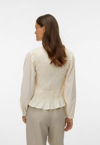 Krēmkrāsas blūze ar smocked aizmuguri, garām pūkainām piedurknēm un volāniem apakšmalā. Izgatavota no viegla, strukturēta auduma.