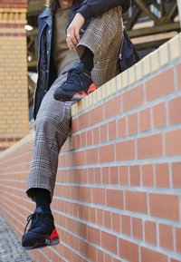 Sneaker neri con dettagli rossi, superiore testurizzato e suola robusta. Pantaloni a scacchi bianchi e neri, seduto contro un muro di mattoni.