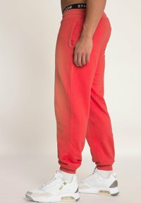 Rode sweatpants met een taps toelopende pasvorm, zijzakken en geribbelde boorden. Draagt een paar witte sneakers met grijze en zwarte accenten.