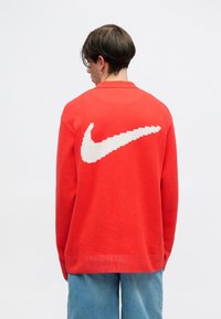 Crvena pletena dukserica s velikim bijelim pikseliziranim Nike logom na leđima, rebrasti ovratnik, rub i manžetne, u kombinaciji s trapericama.