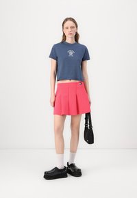 T-shirt cropped bleu marine avec impression graphique, associé à une mini-jupe plissée rose. Chaussures à plateforme noires et chaussettes blanches mi-mollet. Sac bandoulière noir.