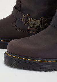 Brown leather ankle boots with a yellow stitched sole, metallic buckle, and branding detail. Textured surface with a slightly distressed finish.