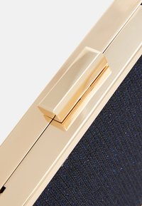 Tiivis clutch-laukku tummansinisen kankaan kanssa, jossa on teksturoitu pinta, ja kiiltävä kultametalinen solki ja yksityiskohdat. Ominaisuudet puhtaat linjat.