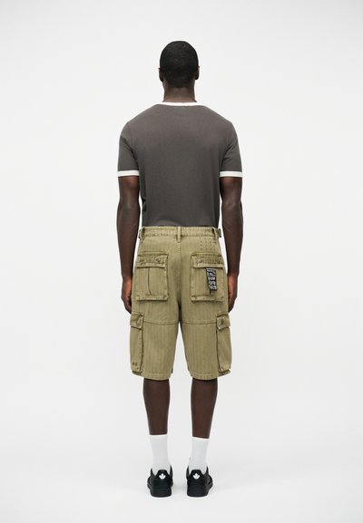 Άνδρας που κοιτάζει προς τα πίσω, φοράει γκρι κοντομάνικο πουκάμισο με λευκή επένδυση και χακί cargo σορτς με πολλαπλές τσέπες, καθώς και μαύρα αθλητικά παπούτσια.