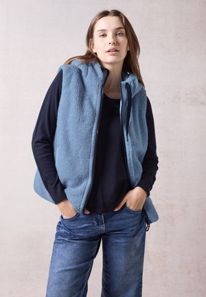 Fleece-Weste in Hellblau mit Reißverschluss, Kapuze und Taschen, getragen über einem navyfarbenen Oberteil, kombiniert mit blauen Jeans. Sanfte Textur.