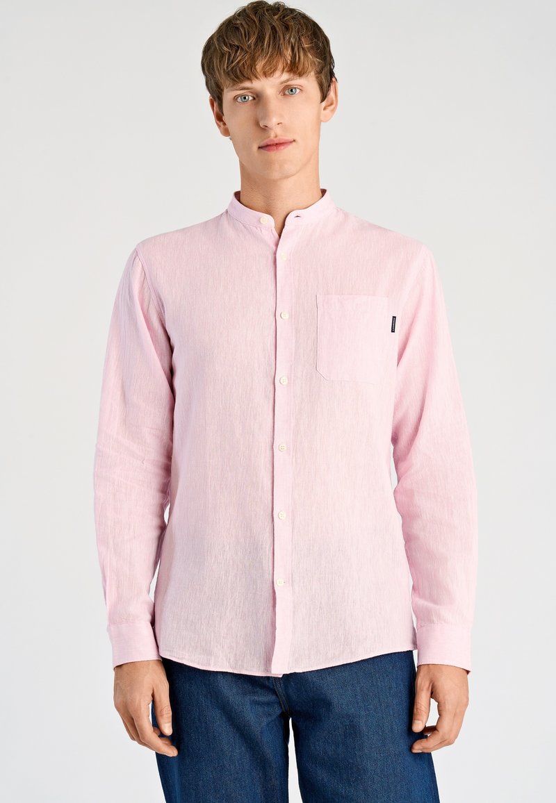 Lichtroze overhemd met lange mouwen, een opstaande kraag, een borstzak aan de voorkant en een subtiele textuur. Knopen aan de voorkant; gecombineerd met donkerblauwe spijkerbroek.