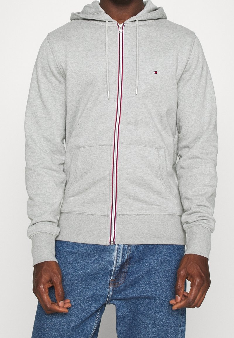 Homme portant un sweat à capuche gris clair zippé avec une fermeture éclair rayée rouge, blanche et bleue et un petit logo sur la poitrine, associé à un jean bleu.