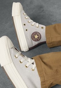 Bež visoke Converse teniske z belimi vezalkami in zlatimi očesci, oblečene z rjavimi hlačami na sivi površini.