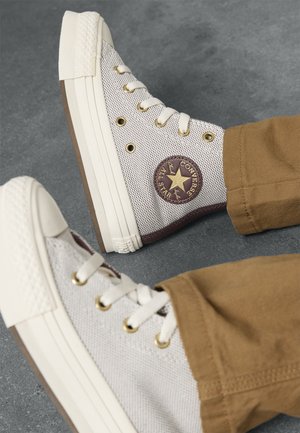 Sneakers beige alte Converse con lacci bianchi e occhielli dorati indossati con pantaloni marroni su un pavimento grigio.