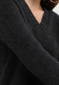 Dunkelgrauer gestrickter Pullover mit V-Ausschnitt, der aus einem weichen, strukturierten Material besteht und über gerippte Details entlang des Ausschnitts und der Ärmelbündchen verfügt.