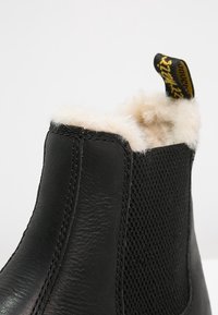 Bottine noire avec cuir lisse, tissu texturé et doublure douce couleur crème. Comprend une languette de tirage noire et jaune.