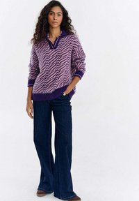 Kobieta z kręconymi włosami, ubrana w fioletowy sweter w wzory, ciemnoniebieskie dżinsy o szerokich nogawkach, brązowe buty i koralikowy naszyjnik, stojąca z jedną ręką w kieszeni.