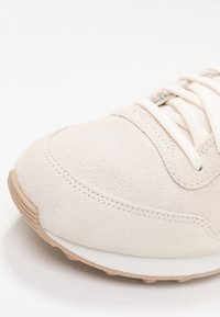 Beige mocka sneakers med vit mellansula och ljusbrun gummisula. Har snörning och synliga sömdetaljer för textur.