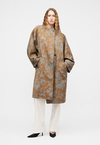 Woman with long wavy hair wearing a loose brown coat with blue floral patterns, white pants, and black pointed heels on a plain white background.