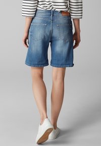 Shorts de mezclilla en un lavado azul claro, con corte recto, dobladillo deshilachado y una etiqueta de marca de cuero marrón en la cintura trasera. Zapatillas blancas.