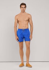 Blauwe zwemshorts met een elastische tailleband en trekkoord, met een gladde textuur en korte lengte, gecombineerd met tan kleurige schoeisel.