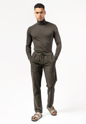 Mann trägt einen dunkelbraunen Rollkragenpullover, passende braune Hose mit Kordelzug und hellbraune Sandalen, steht mit den Händen in den Taschen vor weißem Hintergrund.