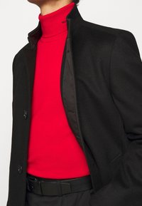 Czarny wełniany płaszcz w połączeniu z włóknami syntetycznymi, z wysokim kołnierzem, pod spodem czerwony golf. Smukły design podkreśla warstwowe tekstury.