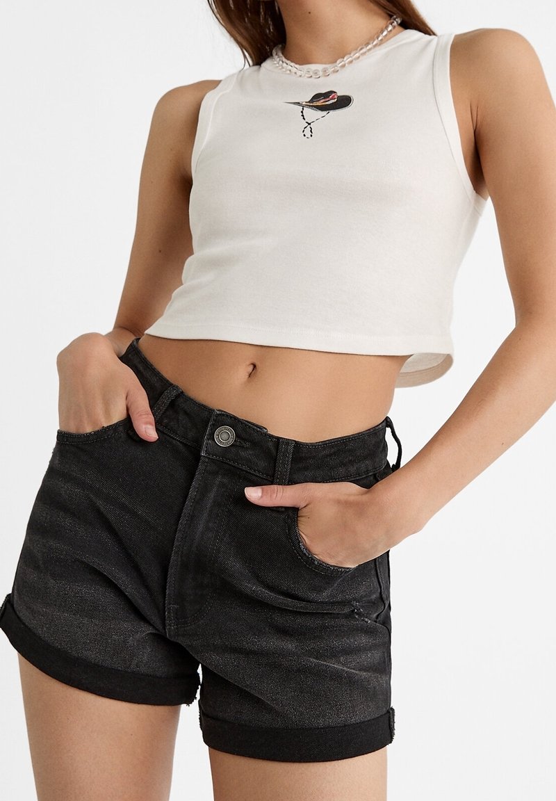 Femme portant un crop top blanc sans manches avec un motif de chapeau et un short en jean noir roulé, les mains dans les poches, sur fond blanc.