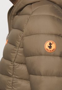 Oliivinvihreä puffer-takki, jossa on tikkikuvio, ja joka näyttää pyöreää oranssia logoa, jossa on ankkamuotoilu, sekä etuvetoketju oranssilla vetonauhalla.