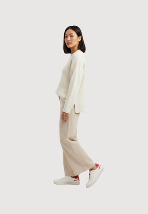Woman with shoulder-length black hair standing sideways wearing a cream ribbed sweater, beige wide-leg pants, and white sneakers with red details.