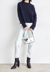 Femme portant un pull bleu marine, un jean bleu clair, des bottes à talons noires, tenant un petit sac à main argenté réfléchissant avec une bandoulière noire.