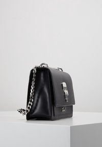 Czarna skórzana torebka z akcentami ze srebrnego metalu, z zamknięciem na klapkę, łańcuszkowym paskiem i charakterystycznym emblematem projektanta z przodu.