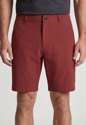 Man draagt roestrode op maat gemaakte korte broek en een donkergrijs ingestopt overhemd, staat met ontspannen armen langs het lichaam.