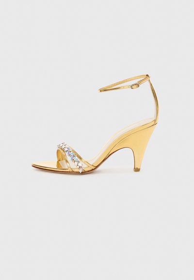 Gold heeled sandal with a strappy design, featuring a rhinestone-accented band. Open toe, adjustable ankle strap, and a tapered heel.