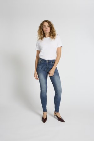 Frau mit lockigem Haar, die ein weißes T-Shirt, blaue Skinny-Jeans und braune High-Heels trägt, stehend vor einem einfarbigen grauen Hintergrund.