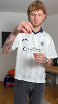 Chemise blanche à manches courtes avec des accents bleu marine et une texture à motifs, jean en denim gris, et une personne tenant un chapelet de perles noires.