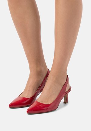 Pies con zapatos de tacón alto slingback de punta puntiaguda y color rojo brillante, con tacones de bloque de altura media sobre un fondo liso.