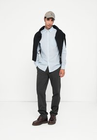 Long-sleeve light blue striped shirt paired with dark gray pants. A black sweater draped over the shoulders and a beige cap complete the look.