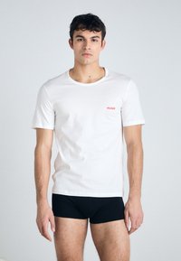 T-shirt en coton blanc à manches courtes, col rond, et petit logo rouge "HUGO" sur la poitrine gauche. Porté avec un boxer noir.