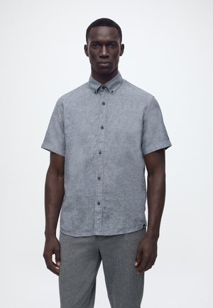 Άνδρας που φοράει γκρι κοντομάνικο πουκάμισο με κουμπιά και γκρι παντελόνι, στέκεται μπροστά σε απλό φωτεινό φόντο.